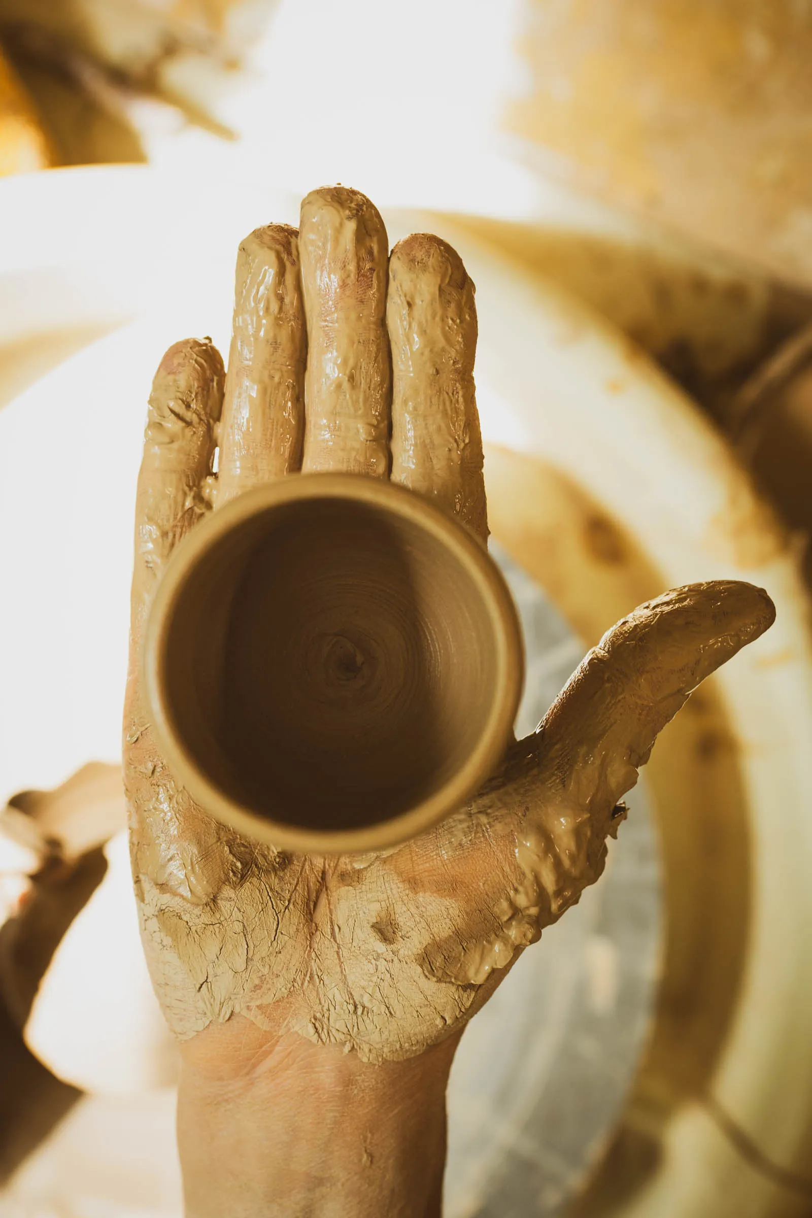 A clay-covered hand holding a freshly thrown cup above the pottery wheel.